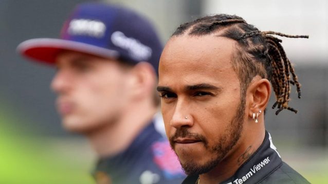 Lewis Hamilton looks down the barrel of the lens with a blurry Max Verstappen looking on in the background