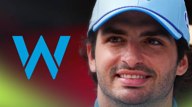 Carlos Sainz wears a blue hat as he smiles with a prominent Williams logo alongside him
