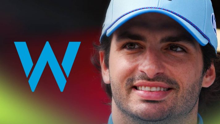 Carlos Sainz wears a blue hat as he smiles with a prominent Williams logo alongside him