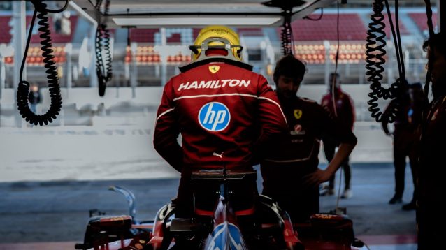 Lewis Hamilton stands in the cockpit of his Ferrari in the garage with his surname visible on the back of his overalls