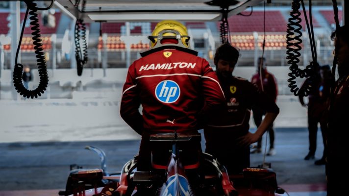 Lewis Hamilton stands in the cockpit of his Ferrari in the garage with his surname visible on the back of his overalls