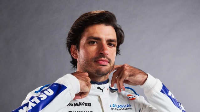 Carlos Sainz holds his hands to his collar as he poses wearing Williams overalls