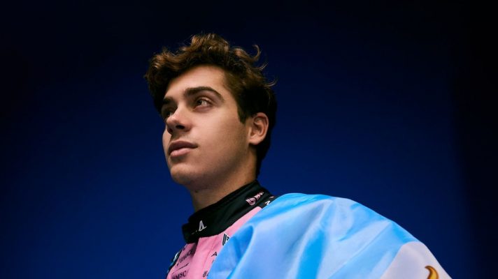 Franco Colapinto poses in Alpine overalls with the flag of Argentina draped over his shoulder