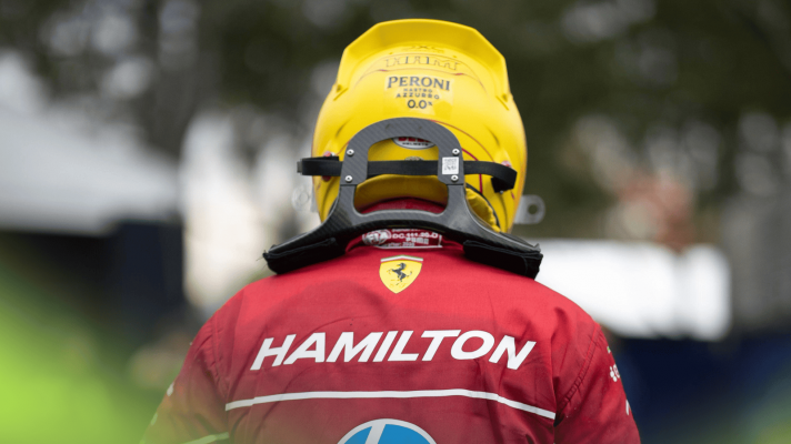 A rear-facing shot of Lewis Hamilton walking through the paddock with his helmet on and his surname written in large letters on the back of his Ferrari overalls