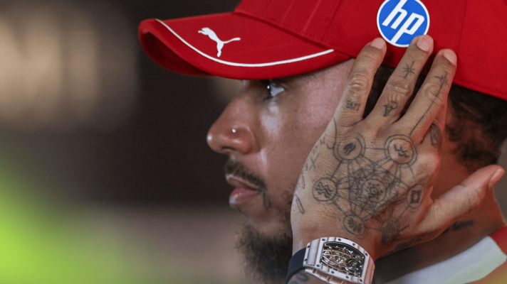 A side-profile shot of Lewis Hamilton holding his hand to his head while wearing a red Ferrari cap