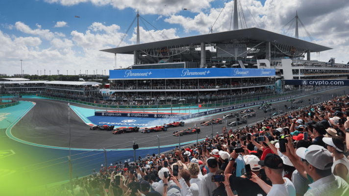 The F1 cars navigate the first corner at the 2024 Miami Grand Prix as fans watch from the grandstands with the stadium in the background