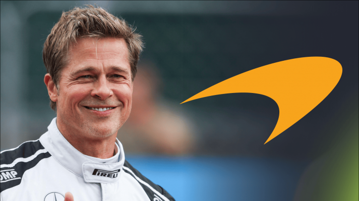 Brad Pitt smiles while wearing racing overalls with an orange McLaren logo alongside him