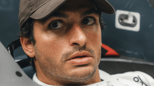 A close-up shot of Carlos Sainz wearing a brown cap and plain-white race suit while carrying out a seat fit