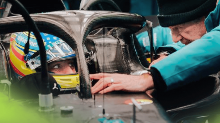 Fernando Alonso glances to his left with his visor open as Adrian Newey leans against the Aston Martin AMR26