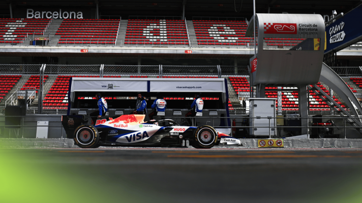A side-profile shot of Liam Lawson's car heading down the Barcelona pit lane with an empty grandstand in the background
