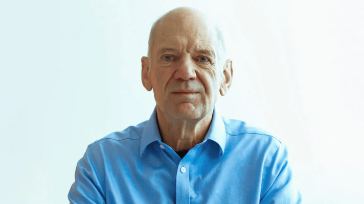 Adrian Newey eyes the camera while wearing a blue shirt with a white background
