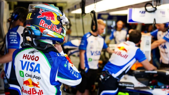 Arvid Lindblad fits his helmet at the front of the Racing Bulls garage as mechanics prepare his car in the background.