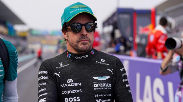 A serious looking Fernando Alonso walks down pit lane with his overalls around his waist.