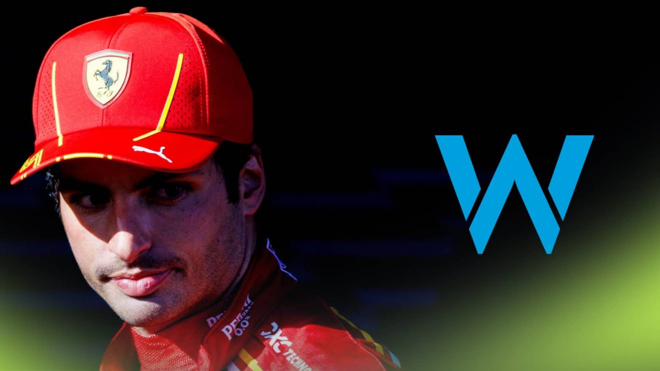 Carlos Sainz stands side-on with a prominent Williams logo alongside him