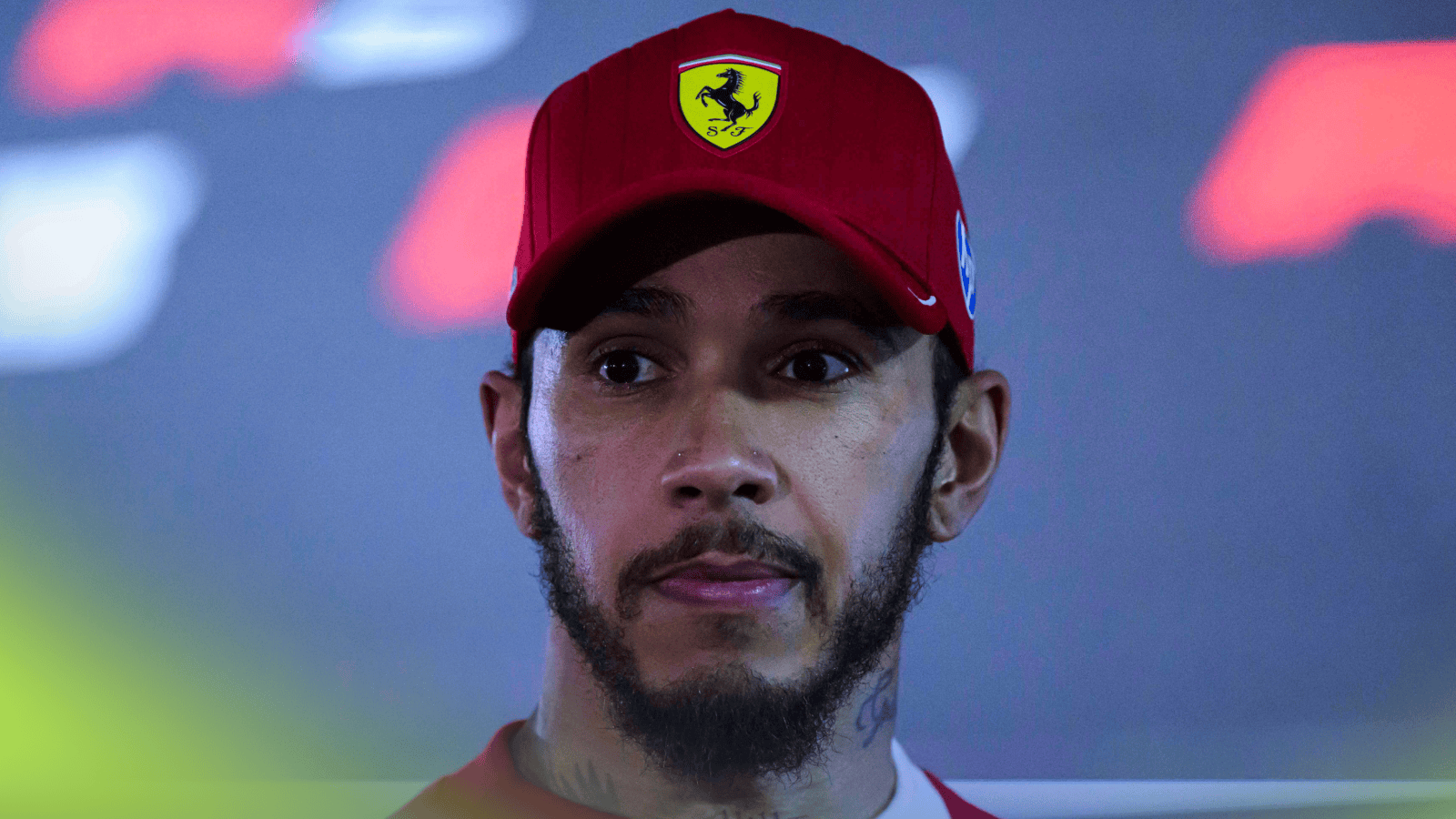 Lewis Hamilton looks tense as he faces the media while wearing a red Ferrari cap