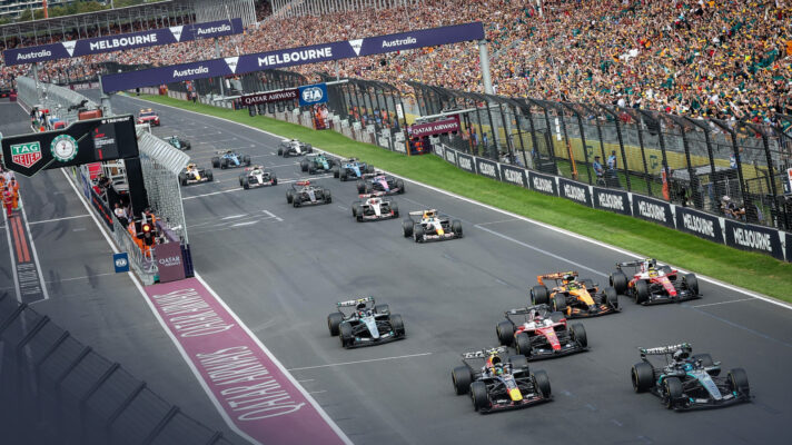 A high shot of the start at the Australian Grand Prix as the pack races away from the line in front of a packed grandstand.