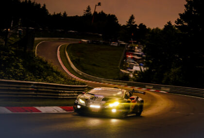 A Lamborghini GT3 car takes a corner on a Nurburgring at dusk, with its headlights blazing.