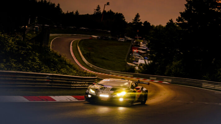 A Lamborghini GT3 car takes a corner on a Nurburgring at dusk, with its headlights blazing.