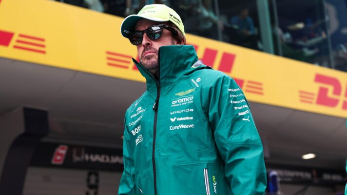 Fernando Alonso in an Aston Martin jacket and sunglasses walks down the pit lane.