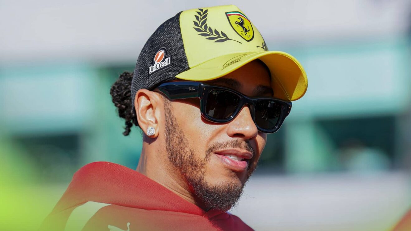 Lewis Hamilton smiles while wearing sunglasses, a red Ferrari hoodie and a yellow cap