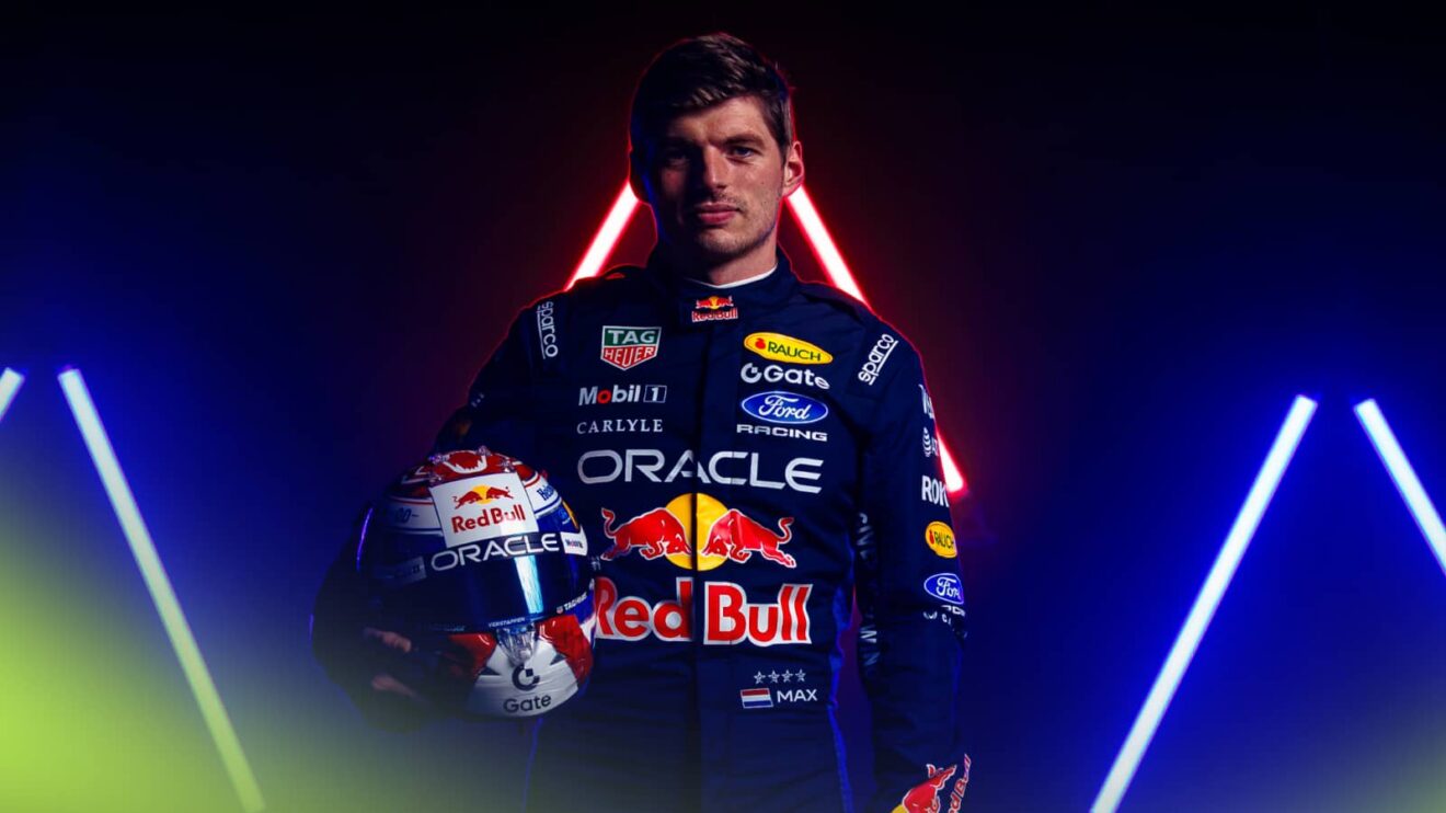 Max Verstappen poses with his helmet while wearing 2026 Red Bull overalls against a dark background