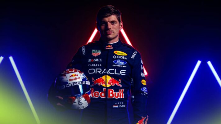 Max Verstappen poses with his helmet while wearing 2026 Red Bull overalls against a dark background