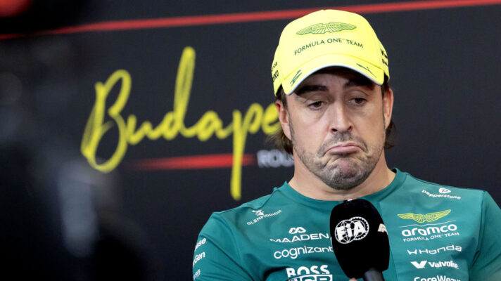 Fernando Alonso, wearing an Aston Martin cap, pulls a face as he holds the mic during a press conference