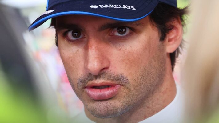 A close-up shot of Carlos Sainz eyeing the camera directly while wearing a navy Williams cap