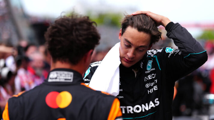 George Russell with his hand on his head and a towel around his neck, Lando Norris blurred in the foreground