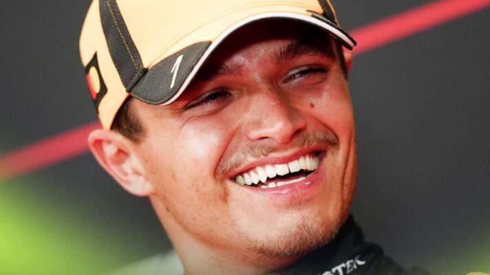 A close-up shot of Lando Norris smiling broadly in a press conference with a McLaren cap on