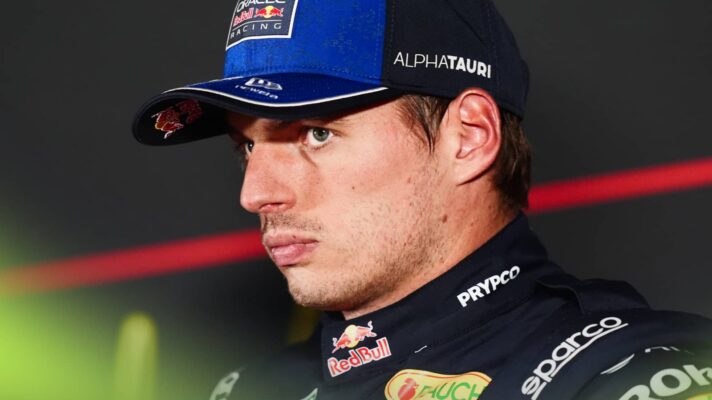 Max Verstappen looks on in a press conference while wearing navy Red Bull overalls and a team cap