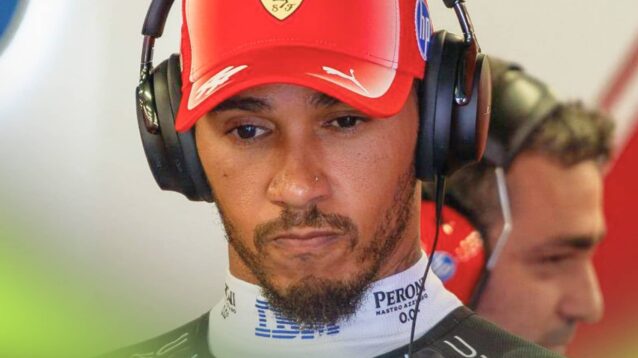 Lewis Hamilton scrunches his face while wearing headphones and a red Ferrari cap in the garage