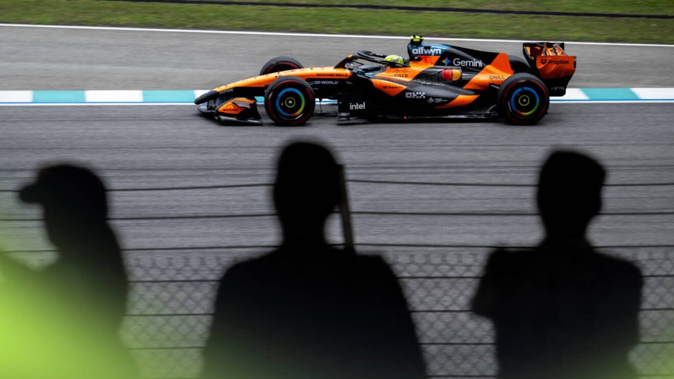 Lando Norris's McLaren on track in Sepang with silhouettes of three fans watching from a grandstand in the foreground