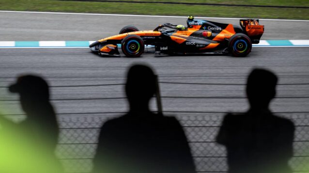 Lando Norris's McLaren on track in Sepang with silhouettes of three fans watching from a grandstand in the foreground