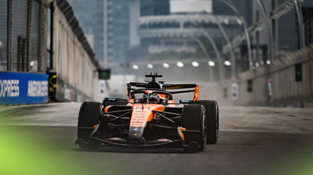 Oscar Piastri's McLaren on track in Singapore during daylight with part of the city's landscape in the background