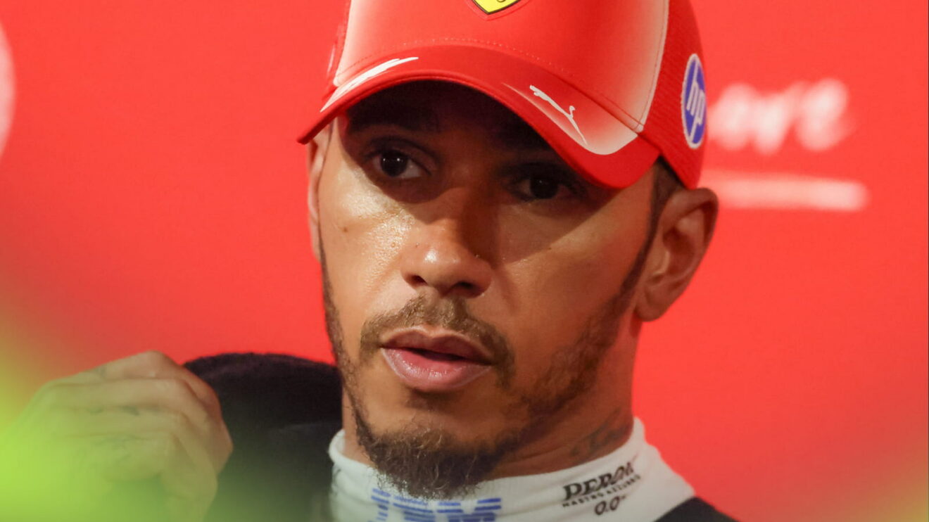Lewis Hamilton wearing a red cap with a towel over his shoulder and bright-red Ferrari branding in the background